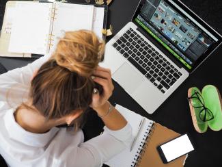 Top-down image of a woman sitting at a cluttered desk. She's holding her head and looking down.